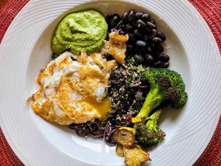 Healthy vegetarian black rice noodle bowl with fried egg, pesto sauce, black beans, and roasted broccoli on a red tablecloth.