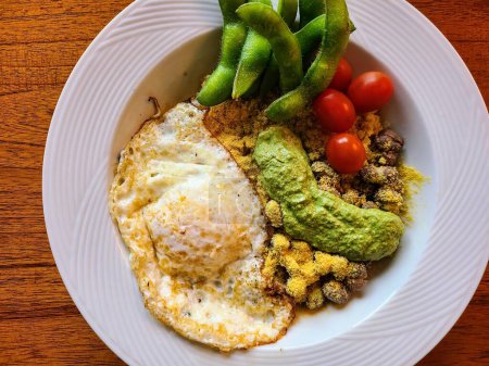 Nutritious vegetarian rice bowl featuring a sunny-side-up egg, pesto sauce, cherry tomatoes, edamame, and nutritional yeast over rice in a white bowl on a wooden table.
