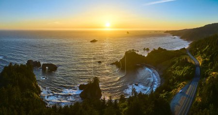 Aerial view of Arch Rock in Brookings, Oregon, capturing the winding coastal road and dense green forests at sunset. The golden hour light enhances the tranquility and natural beauty of the Samuel H