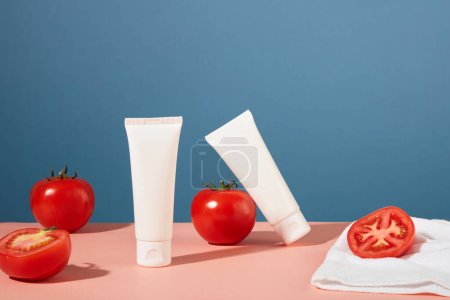 Téléchargez les photos : Vue de face de deux tubes cosmétiques non étiquetés sont placés sur une table rose avec des tomates fraîches. Concept cosmétique avec des ingrédients naturels d'extrait de tomate. - en image libre de droit