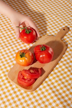 Foto de La mano sostiene un jugoso tomate rojo colocado en una tabla de cortar de madera con otros tomates. Mantel a cuadros con dos colores blanco y amarillo. Espacio creativo. - Imagen libre de derechos