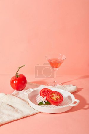 Téléchargez les photos : Des tomates tranchées et une fleur de camélia sont placées sur un plateau rond en céramique, à côté d'une tomate et d'un verre à cocktail contenant du jus de tomate sur fond rose. Espace de copie pour annonces. - en image libre de droit