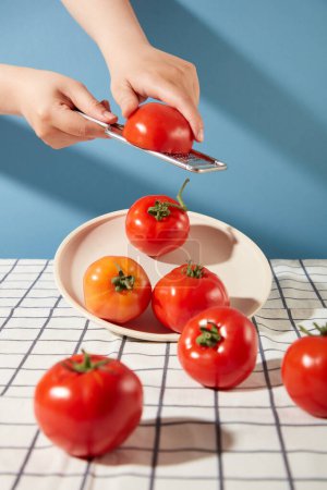 Photo for Front view of a hand holds a vegetable grater, the other holds half a tomato. On the table are red tomatoes with a plaid tablecloth and a blue background. - Royalty Free Image