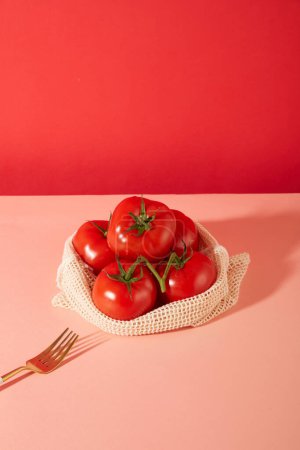 Téléchargez les photos : Les tomates rouges juteuses sont stockées dans un panier en tissu de maille, à côté d'une fourchette en métal sur une table rose pastel avec un fond rose foncé. Espace de copie pour la publicité. - en image libre de droit