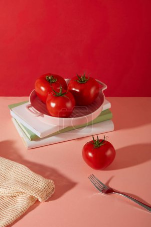 Foto de Tres tomates se colocan en una bandeja de cerámica rosa en la parte superior de los cuadernos. Un tomate se muestra en una mesa rosa con un tenedor de metal. Los tomates contienen muchas vitaminas que son buenas para la salud. - Imagen libre de derechos