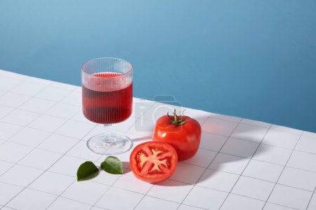 Téléchargez les photos : Un verre contenant un liquide rouge transparent est placé à côté de tomates fraîches et de feuilles vertes sur un fond bleu et blanc. Les tomates contiennent de nombreuses vitamines B telles que B1, B3, B5, B6, B9. - en image libre de droit