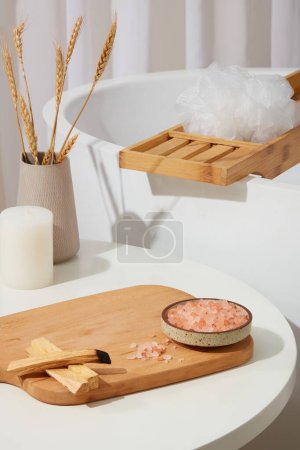 Photo for Wooden chopping board featured a dish of pink himalayan salt and some palo santo wood smoke. Bathroom concept with a bathtub behind. Empty space for product presentation - Royalty Free Image