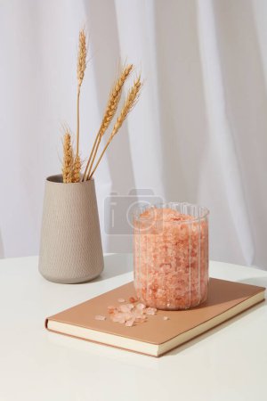 Photo for A jar containing a lot of pink himalayan salt placed on a notebook, displayed with flower pot. White curtain decorated. Himalayan salt can benefit even the most sensitive skin types - Royalty Free Image