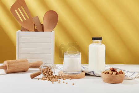 Photo for Front view of a milk cup placed on wooden podium, displayed with a bowl of nuts, soybeans, kitchen utensil and a milk bottle. Empty label bottle for product promotion - Royalty Free Image