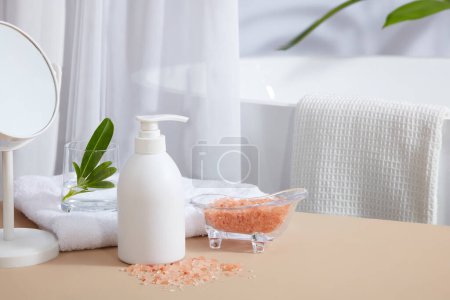 Téléchargez les photos : Concept de salle de bain avec une étiquette vide bouteille blanche décorée d'une baignoire en verre contenant du sel himalayen rose et quelques fournitures. Maquette de soin de la peau bouteille cosmétique de beauté faciale - en image libre de droit