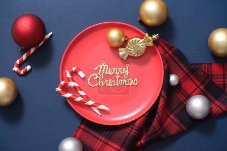 Téléchargez les photos : Vue de dessus de la plaque de céramique rouge se détachant sur le fond bleu, décorée avec du tissu à carreaux, des boules colorées et des cannes à bonbons. Joyeux vœux de Noël au centre du cadre - en image libre de droit