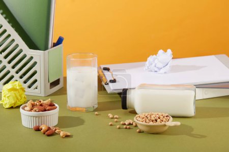 Photo for An office desk featured some office supplies with a cup and bottle of milk. Two bowls containing soybeans, cashew nuts and almond. Blank label bottle for mockup - Royalty Free Image