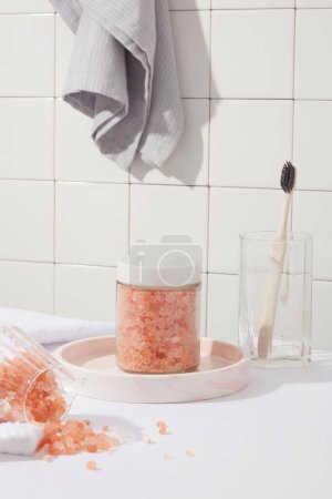 Foto de Sal rosada del himalaya contenida dentro del frasco de etiqueta en blanco. Concepto de baño con una toalla colgada en la pared. La sal rosada del himalaya puede contener trazas de nutrientes y minerales - Imagen libre de derechos
