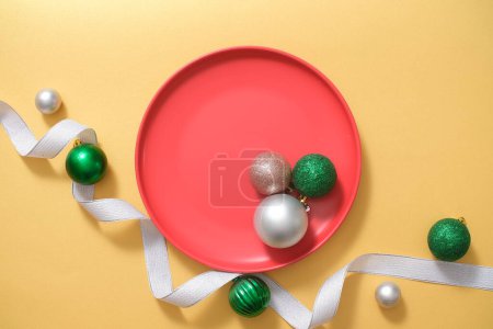 Foto de Composición del marco de Navidad o Año Nuevo con placa de cerámica roja mostrada sobre fondo amarillo con bolas plateadas y verdes. Espacio en blanco para el producto de visualización. Vista superior, plano - Imagen libre de derechos
