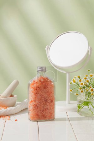 Foto de Botella de vidrio que contiene una gran cantidad de sal himalaya rosa arreglada con un espejo y una maceta de flores de margarita. Hay varias afirmaciones sobre los beneficios para la salud asociados con el consumo de sal rosa - Imagen libre de derechos