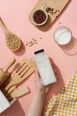 Foto de Utensilios de cocina, soja, frijoles rojos y una taza de leche que se muestra en la superficie de color rosa. Una mano sosteniendo una botella de leche sin etiquetar. Etiqueta vacía para la maqueta del producto de bebidas - Imagen libre de derechos