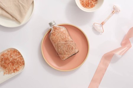 Téléchargez les photos : Une bouteille contenant du sel d'himalaya rose placée sur un plat rose, entourée d'un ruban, d'un rouleau de massage facial et d'une serviette blanche. Concept de produits de beauté naturels - en image libre de droit