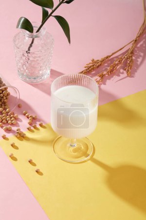Foto de Un vaso de leche de soja colocado en la superficie rosa y amarilla con soja y una olla de hojas verdes. La leche de soja promueve niveles de energía saludables con proteínas y vitaminas B - Imagen libre de derechos