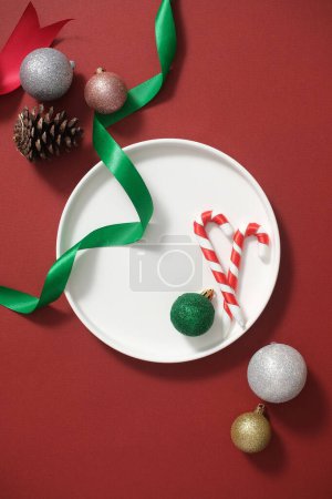 Foto de Vista superior de plato de cerámica blanca decorada con bolas de colores, bastones de caramelo y cinta verde sobre un fondo rojo. Espacio en blanco en el plato para el producto de exhibición. concepto de vacaciones de Navidad - Imagen libre de derechos