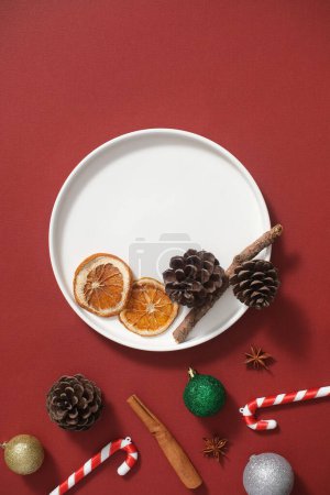 Foto de Fondo creativo con el concepto de vacaciones de Navidad para la publicidad. En el fondo rojo, plato redondo blanco decorado con cono de pino seco, palo de canela, bastón de caramelo y bolas de colores - Imagen libre de derechos