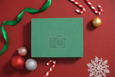 Téléchargez les photos : Vue de dessus d'une boîte cadeau verte affichée sur fond rouge avec des boules colorées, des cannes à bonbons, un ruban et des flocons de neige décorés. Espace vide pour le produit d'affichage - en image libre de droit