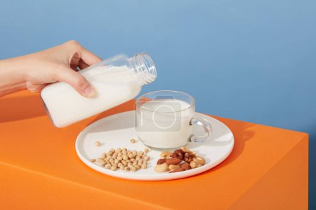 Foto de Un plato en forma redonda que contiene soja, almendras y anacardos con una taza de leche. Modelo de mano sosteniendo una botella llena de leche de nuez. Etiqueta en blanco para la maqueta del producto - Imagen libre de derechos