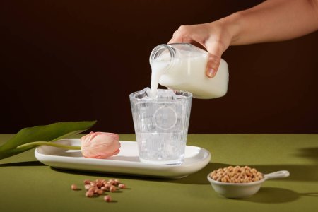 Foto de Una mano está vertiendo leche de una botella en una taza con hielo dentro. Soja contenida en un tazón y algunos cacahuetes exhibidos. La leche de nuez tiene muchas ventajas para la salud - Imagen libre de derechos