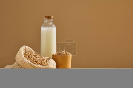 Téléchargez les photos : Une bouteille en verre remplie de lait de soja est décorée d'un filet et d'un bol en bois de graines de soja. Espace pour la présentation de produits cosmétiques ou d'emballages - en image libre de droit