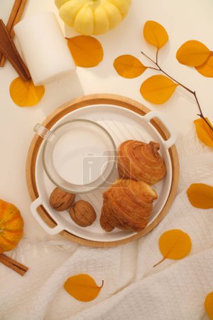 Foto de Foto de un aperitivo con concepto cálido de otoño. En la bandeja hay deliciosos croissants y un vaso de leche caliente. Destacan las hojas amarillas secas, decoradas con calabazas y velas blancas - Imagen libre de derechos
