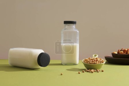 Téléchargez les photos : Deux bouteilles vierges de lait de soja décorées sur une surface verte avec un plat de graines de soja. Plat en bois contenant quelques noix. Le soja est un ingrédient très spécial - en image libre de droit