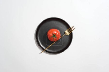 Foto de Plato negro en forma redonda contó con un tomate y tenedor de oro. Fondo blanco. El tomate (Solanum lycopersicum) también es una fuente de nutrientes - Imagen libre de derechos