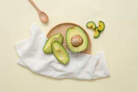 Photo for Creative cosmetics flat lay with ingredient from avocado. Top view of fresh avocado slices decorated with white cloth with wooden dish and spoon on beige background. Natural cosmetics concept. - Royalty Free Image