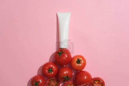 Téléchargez les photos : Tube cosmétique vierge de couleur blanche posé sur un tas de tomates. Fond rose. Les tomates regorgent de vitamines, de nutriments et de minéraux - en image libre de droit