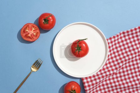 Foto de Un tomate colocado en plato de cerámica en forma redonda, decorado con un tenedor y tela a cuadros rojo y blanco. Los tomates (Solanum lycopersicum) son una excelente fuente de vitamina C - Imagen libre de derechos