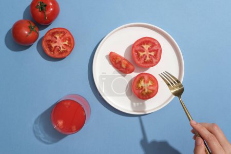 Foto de Un plato blanco presentaba algunas rebanadas de tomate decoradas con un vaso de jugo de tomate. Modelo de mano sosteniendo un tenedor. Los tomates son ricos en compuestos antiinflamatorios - Imagen libre de derechos