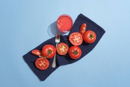 Téléchargez les photos : Tomate coupée en deux et les tranches sont placées sur un tissu avec une fourchette. Un verre de jus de tomate sur fond bleu. Le jus de tomate fournit de nombreux ingrédients sains - en image libre de droit