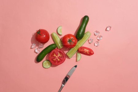 Téléchargez les photos : Concombre et tomate coupés en tranches et moitiés décorées sur fond rose avec des glaçons et un couteau. Concept de fruits sains qui sont bons pour la peau et la santé - en image libre de droit