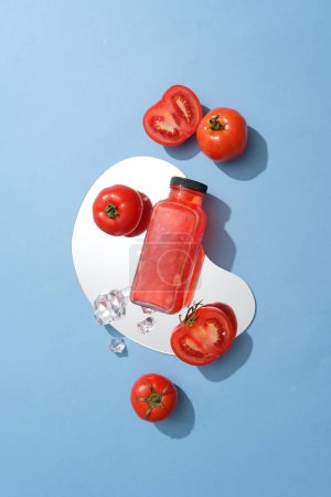 Foto de Botella de vidrio sin etiquetar llena de jugo de tomate colocado en un espejo con tomates y cubitos de hielo. El tomate tiene muchos beneficios cuando se aplica a la piel - Imagen libre de derechos