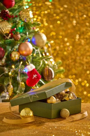 Téléchargez les photos : Un coffret cadeau ouvert rempli de beaucoup de boules à l'intérieur présenté sur une surface jaune étincelante. Arbre de Noël affiché. Alors que les Américains commençaient à embrasser Noël comme des vacances parfaites en famille - en image libre de droit
