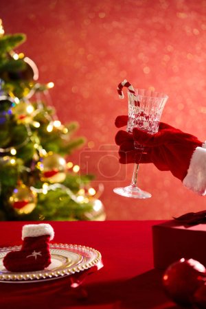 Foto de Modelo de mano con guantes de Santa Claus sosteniendo una copa de vino. Un plato con calcetines de lana. El día de Navidad es celebrado religiosamente por la mayoría de los cristianos - Imagen libre de derechos