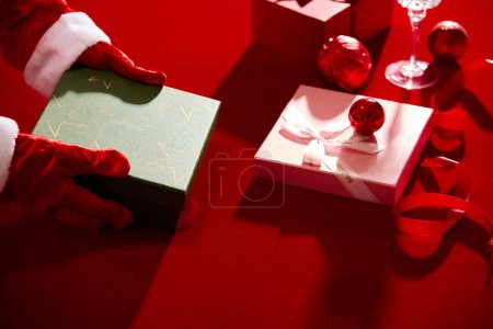 Téléchargez les photos : Coffrets cadeaux, rubans et boules rouges affichés sur la table rouge, un modèle à main avec gants id tenant une boîte. Le jour de Noël est un jour férié dans de nombreux pays - en image libre de droit