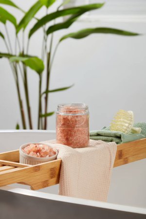 Photo for Wooden bathtub tray featured a glass jar and ceramic bowl of pink himalayan salt and a scalp massage brush. Pink himalayan salt often has larger crystals than table salt - Royalty Free Image