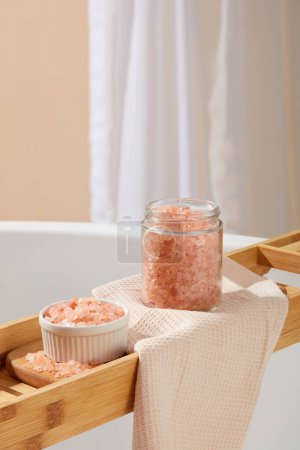 Téléchargez les photos : Vue rapprochée d'un bol et d'un bocal transparent contenant du sel himalayen rose placé sur un plateau de baignoire en bois. Le sel d'himalaya rose est un exfoliant naturel brillant - en image libre de droit