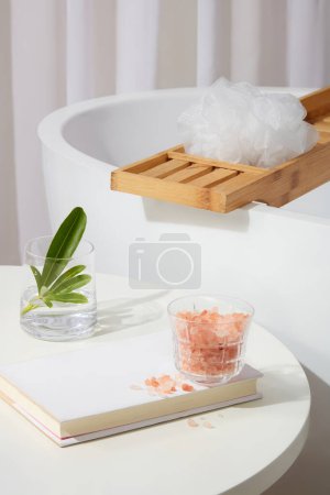 Téléchargez les photos : Une table comportait un livre avec une tasse en verre contenant du sel himalayen rose posé dessus. Baignoire avec un plateau en bois à travers. Le sel d'himalaya rose peut profiter même aux types de peau les plus sensibles - en image libre de droit