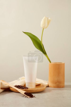 Photo for Front view of white plastic tube without label decorated with coffee beans and coffee powder on wooden dish with vase of flower on white background. Mockup scene for cosmetic. Space for design. - Royalty Free Image