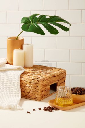 Photo for On the white tile background, coffee beans on wooden tray decorated with towel, candle and green monstera leaves. Blank space on basket for display bath products - Royalty Free Image