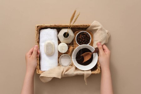 Foto de Dos manos de mujer sosteniendo una cesta que contiene objetos para bañarse, granos de café y café en polvo, sal de baño, vela y toalla sobre fondo marrón claro - Imagen libre de derechos