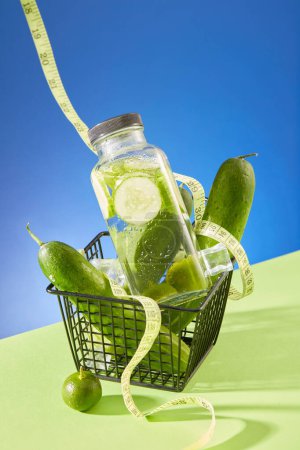 Photo for A basket containing cucumbers and a glass bottle filled with cucumber detox water. A tape measure displayed. Empty label bottle for branding mockup design - Royalty Free Image