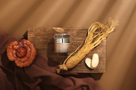 Auf einem Holzpodest steht ein Kosmetikgefäß, das mit Wurzel und Scheiben Ginseng, Lingzhi-Pilz und braunem Tuch auf braunem Hintergrund dekoriert ist. Konzept für Kosmetika aus seltenen Kräutern. Ansicht von oben