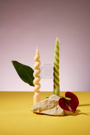 Foto de Dos velas retorcidas en dos colores: beige y verde, una losa de piedra, hojas verdes y flores se muestran en una mesa amarilla, fondo púrpura claro. Espacio vacío para la visualización del producto. - Imagen libre de derechos